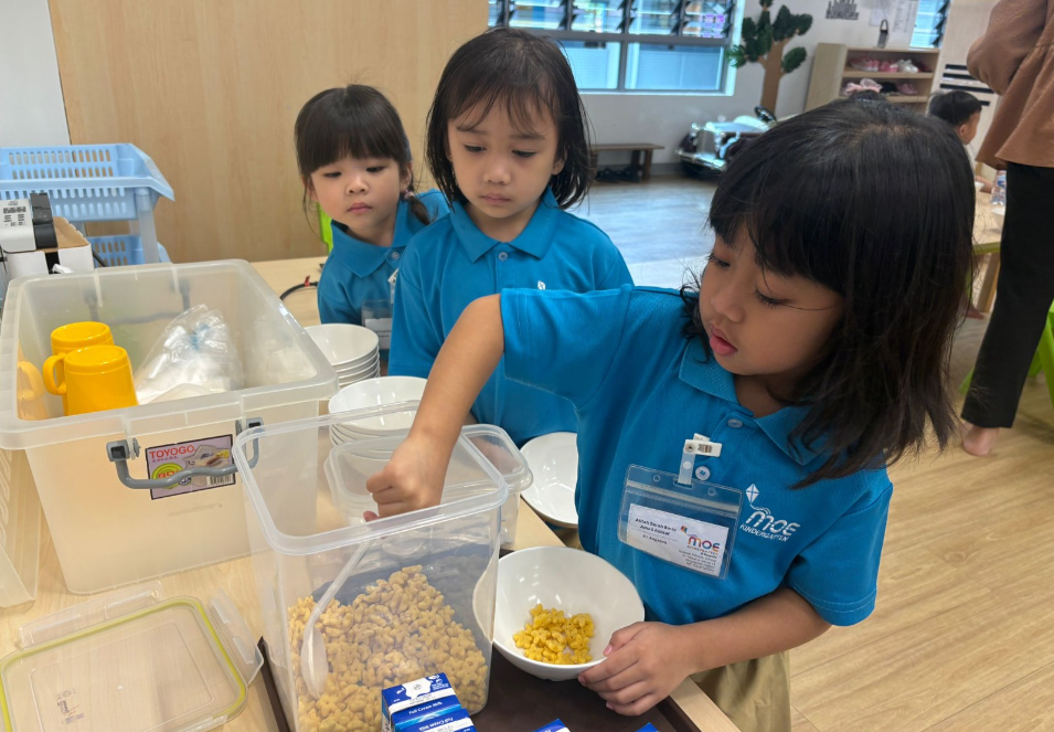 Girl in blue polo scoops cereal into a bowl while two children watch beside bins of dishes and cups.